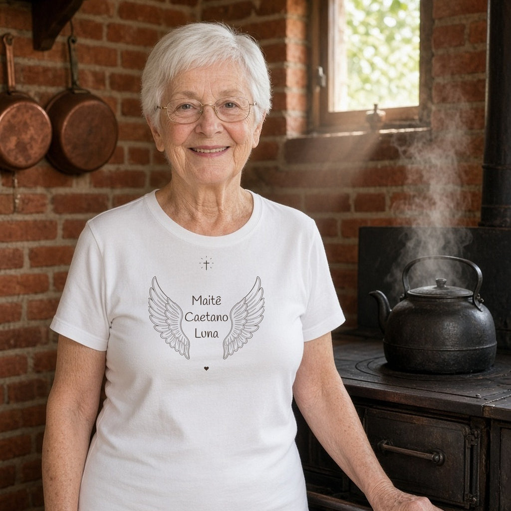 Avó de cabelo branco e óculos em uma cozinha com fogão a lenha vestindo a camiseta de algodão peruano Os Anjos da Vovó branca com asas de anjo e os nomes de 3 netos