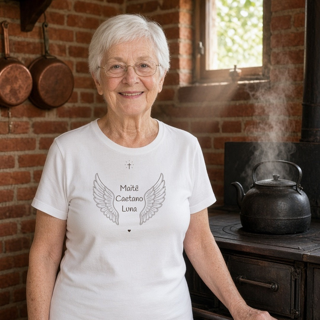 Avó de cabelo branco e óculos em uma cozinha com fogão a lenha vestindo a camiseta de algodão peruano Os Anjos da Vovó branca com asas de anjo e os nomes de 3 netos