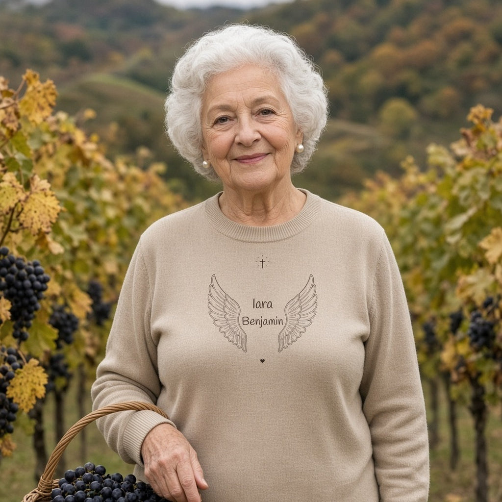 Avó de cabelo branco ondulado em um vinhedo com cesto de uvas vestindo o suéter Os Anjos da Vovó areia com asas de anjo e os nomes de 2 netos